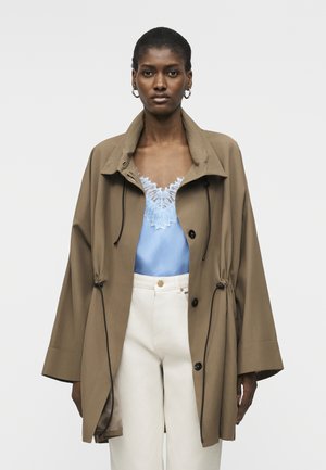 Moteris dėvi laisvą rudos spalvos paltą, po juo šviesiai mėlyną viršutinę dalį su nėrinių apdaila ir baltas aukšto liemens kelnes, stovinti priešais paprastą foną.