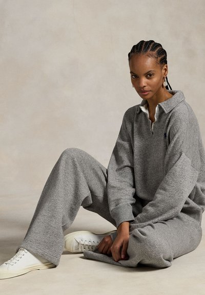 Γυναίκα με πλεξούδες που κάθεται σταυροπόδι, φοράει γκρι φούτερ με φερμουάρ, ταιριαστό φαρδύ παντελόνι, άσπρα αθλητικά παπούτσια και σκουλαρίκια με κοχύλια.