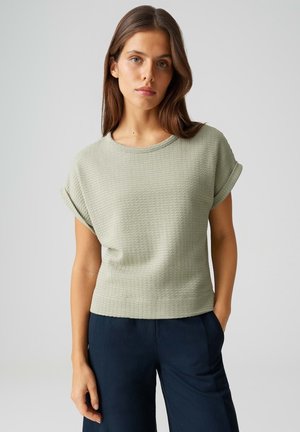 Junge Frau mit glattem braunem Haar, die ein hellgrünes, strukturiertes Kurzarmoberteil und dunkelblaue Hosen trägt, steht mit einer Hand in der Tasche.