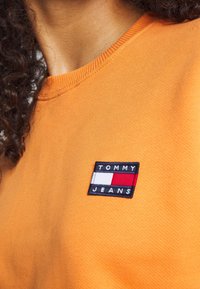 Oranžová bavlněná mikina s kulatým výstřihem a obdélníkovou nášivkou s nápisem "TOMMY JEANS" bílým textem na černém pozadí.