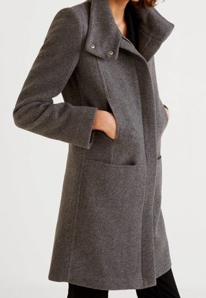 Frau trägt einen knielangen, dunkelgrauen Wollmantel mit hohem Kragen und Händen in den Seitentaschen, abgebildet von den Schultern bis zu den Oberschenkeln.