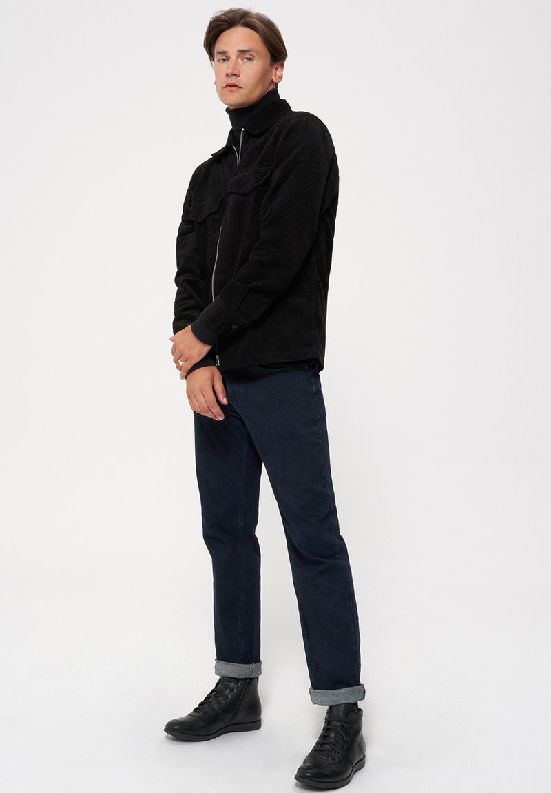 Czarna kurtka kordurowa z wysokim kołnierzem, z kieszeniami z przodu, noszona na czarnym golfie. Ciemnoniebieskie spodnie z podwiniętymi nogawkami i czarne sznurowane botki.