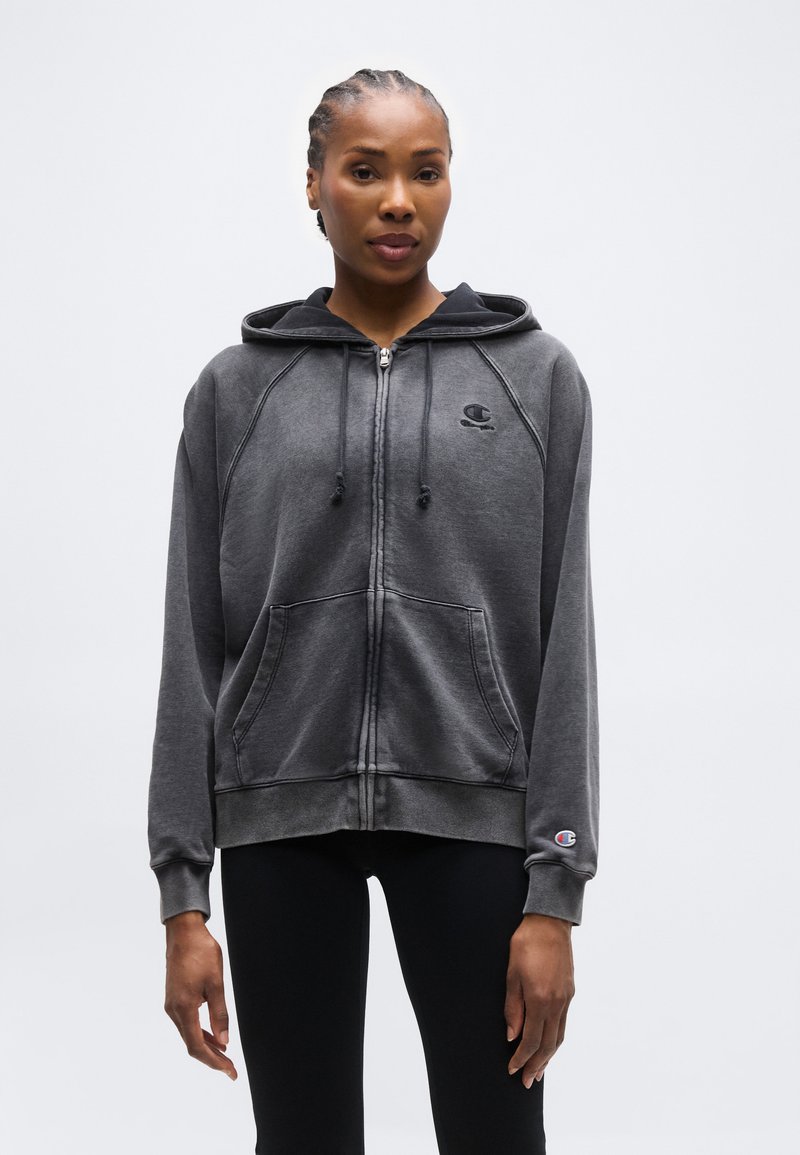 Γυναίκα με πλεξούδες που φοράει σκούρο γκρι ζακέτα με φερμουάρ και τσέπες μπροστά, καθώς και μαύρο κολάν, στέκεται μπροστά σε απλό φόντο.