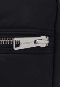 Detaliu al unei genți din țesătură neagră, cu un fermoar argintiu și un maner dreptunghiular, gravat cu "Carhartt" și înconjurat de o textură subtilă.