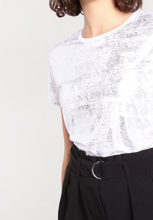 Vrouw met een wit shirt met korte mouwen en een subtiel patroon, ingestopt in een hoge zwarte broek met een riem en metalen gesp.