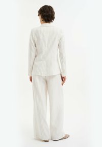 Abito bianco a righe sottili su misura con maniche lunghe, colletto a indicazione e pantaloni a gamba larga. Il materiale sembra leggero e strutturato.