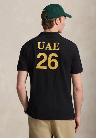 Άνδρας που φοράει μαύρο πόλο με κίτρινο "UAE 26" στην πλάτη, πράσινο καπέλο και μπεζ παντελόνι, στέκεται με την πλάτη στην κάμερα.