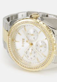 Orologio in metallo dorato e argentato con quadrante bianco, tre sottoquadranti e una lunetta incrustrata di cristalli. Presenta indicatori orari lineari e una corona.
