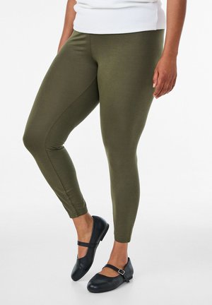 Person wearing olive green leggings and black Mary Jane shoes standing with one leg slightly bent against a white background.