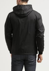 Zwarte lichtgewicht jas met een capuchon, een gladde textuur, elastische manchetten en een relaxte pasvorm, weergegeven vanaf de achterkant met donkere spijkerbroek.