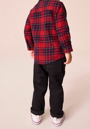 Chemise à carreaux rouge et vert à manches longues, associée à un pantalon noir et des baskets blanches, présentant des poignets retroussés. Vue de dos.