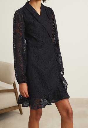 Femme portant une robe portefeuille en dentelle noire avec de longues manches transparentes et un ourlet à volants, debout près d'une chaise beige dans une pièce neutre.