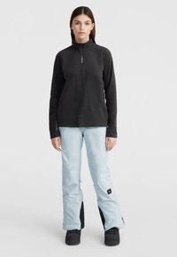 Zwarte fleece pullover met een kwart-rits aan de voorkant, gecombineerd met lichtblauwe sneeuwbroeken met zwarte accenten en een logo-patch, en zwarte laarzen.