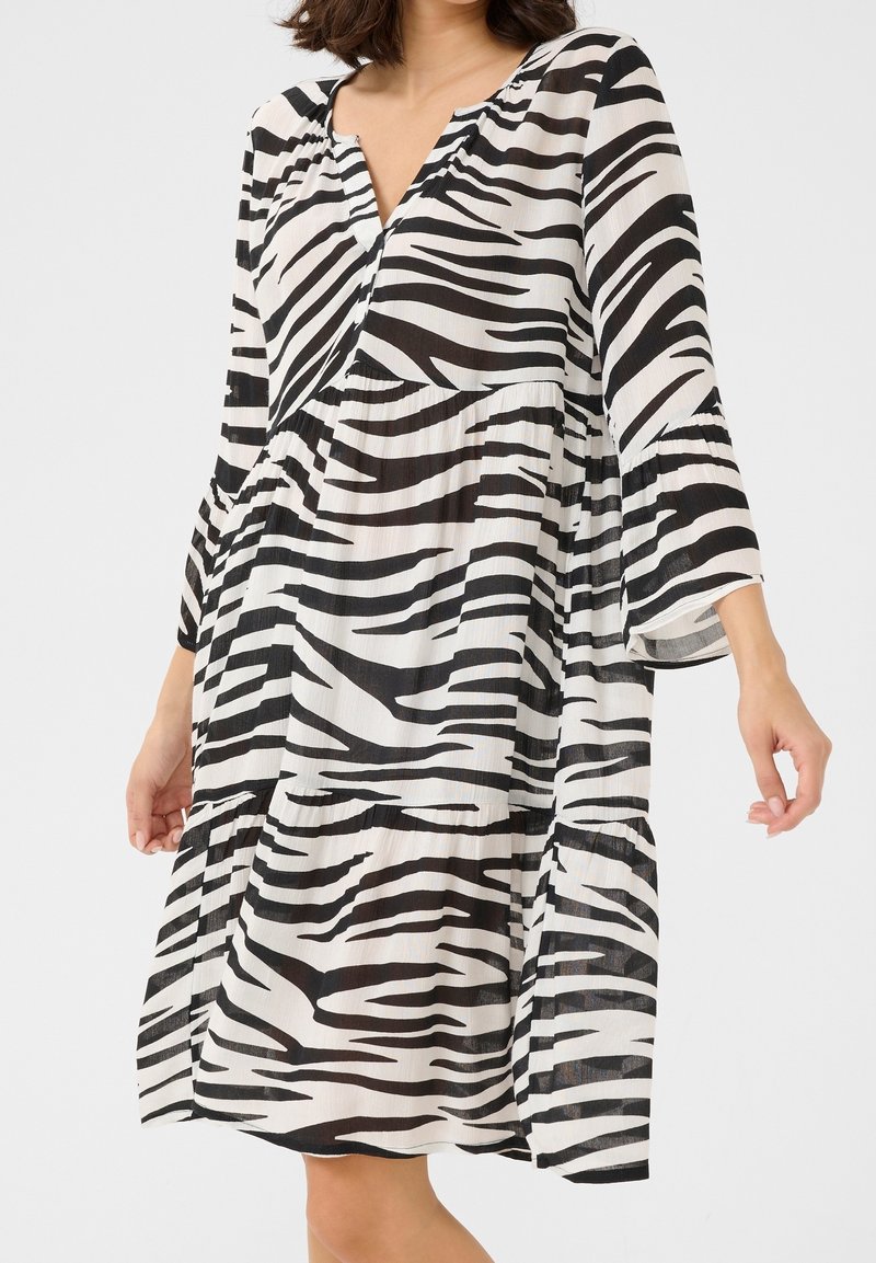 Femme portant une robe ample à imprimé zèbre noir et blanc avec manches 3/4 et col en V, debout devant un fond uni.