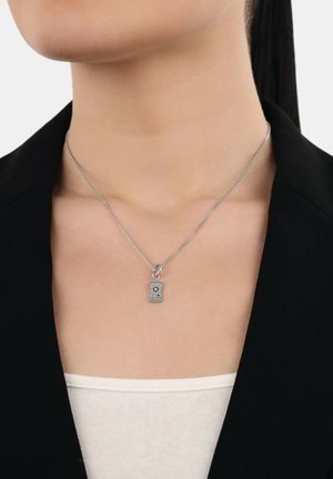 Silberne Kettenhalskette mit einem kleinen rechteckigen Anhänger, der ein Sonnensymbol und Text darstellt, getragen über einem weißen Oberteil und einem schwarzen Blazer.