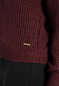 Gros plan sur une personne portant un pull en maille côtelée bordeaux avec une petite étiquette rectangulaire en or et un pantalon noir.