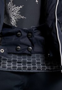 Gros plan d'une veste bleu marine avec des boutons noirs, une fermeture éclair blanche et un tissu intérieur présentant des motifs argentés de flocons de neige et d'hexagones.