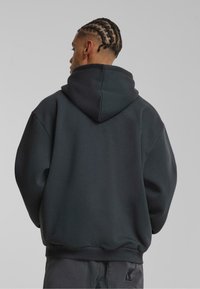Mørkegrå hættetrøje med oversized pasform, ribbede manchetter og kant, glat tekstur, set fra bagsiden med et logo nederst til venstre.
