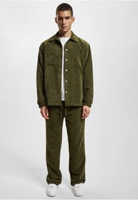 Chaqueta de pana verde con bolsillos en el pecho y botones blancos, combinada con pantalones a juego. Una camiseta blanca y zapatillas deportivas completan el atuendo.