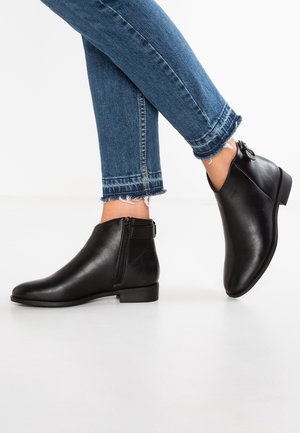 Person iført sorte læderankelstøvler med sidelås og blå slidte skinny jeans med frynser mod hvid baggrund.