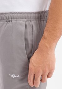 Lichtgrijze trainingsbroek met een elastische tailleband, zijzakken en een klein wit logo op de linkerdij. Gladde stoftextuur.