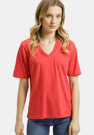 Blondynka z falującymi włosami, ubrana w czerwoną koszulkę z dekoltem w serek i niebieskie jeansy, stojąca na jasnym, jednolitym tle.