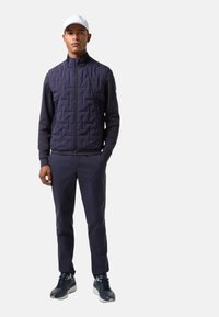 Navy quilted vest with high collar, paired with navy trousers. Model wears white cap and dark athletic shoes. Simple, functional design.