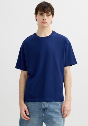 Joven con cabello corto y castaño, vistiendo una camiseta sencilla azul marino de manga corta y vaqueros azul claro, de pie frente a un fondo liso.