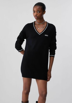 Donna che indossa un vestito maglione nero a maniche lunghe con scollo a V e rifiniture bianche, con collane argento a strati, in piedi davanti a uno sfondo semplice.