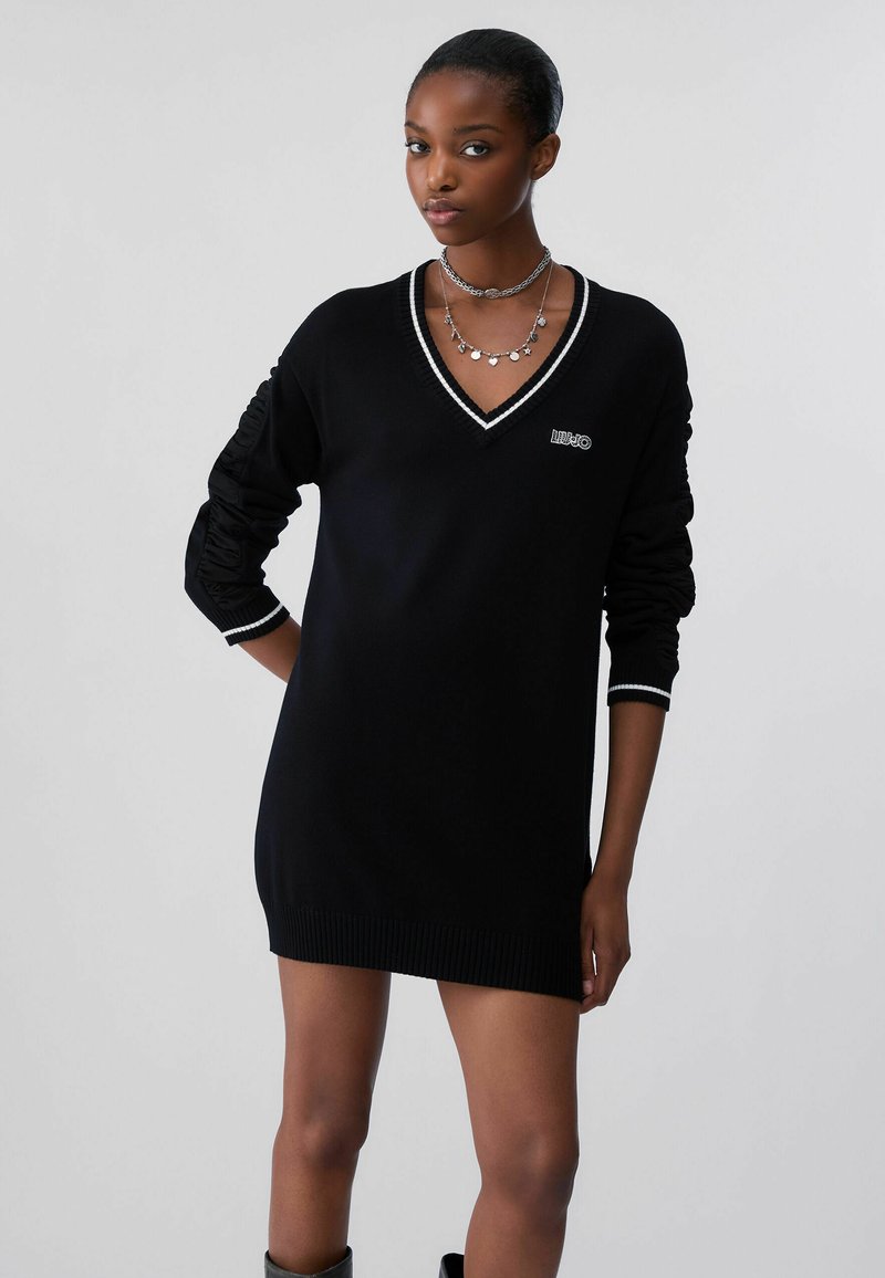 Femme portant une robe pull noire à manches longues, col en V avec une bordure blanche et des colliers argentés superposés, debout devant un fond uni.