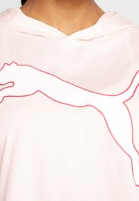 Persoon in een lichtroze hoodie met een groot wit puma-logo omlijnd in het rood over de borst.