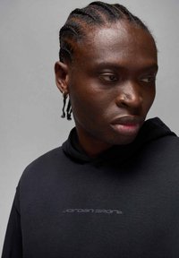 Czarna bluza z kapturem o teksturowanym wykończeniu, z szarym logo "Jordan Sport" na przodzie. Osoba ma zaplecione włosy i małą kolczyk w kółko.