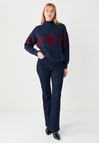 Maglione lavorato a maglia blu navy con motivi a diamante color bordeaux, collo alto e vestibilità ampia. Abbinato a pantaloni svasati in denim scuro e scarpe nere a punta.