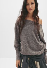 Brown, textured knit sweater with an off-shoulder design and ribbed cuffs. Worn over a layered skirt, showcasing relaxed fit and fabric blend.