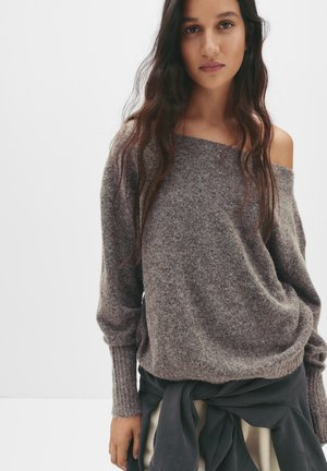 Bruine, gestructureerde gebreide trui met een off-shoulder ontwerp en geribbelde manchetten. Geschreven over een gelaagde rok, die een relaxte pasvorm en stoffenmix tentoonstelt.