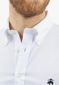 Witte blouse met kraag, voorzien van parelknoopjes en een klein marineblauw logo aan de linkerkant. Gladde, frisse stoftextuur.