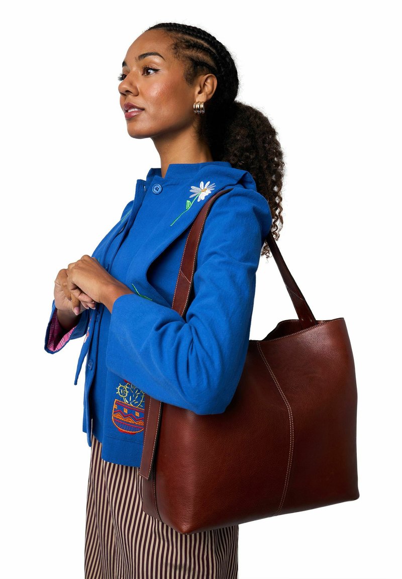 Ruskea nahkainen olkalaukku, jossa on sileä pinta, kaarevat reunat ja kontrastisommitelut. Laukku on kannettu olalla sinisen takin päällä.