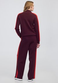 Femme portant un survêtement bordeaux avec des rayures rouges sur les manches et les jambes, se tenant dos à la caméra devant un fond uni.