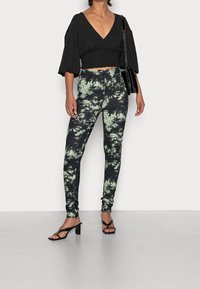 Top corto nero a scollo a V con maniche a palloncino abbinato a leggings tie-dye neri e verdi, e sandali neri con tacco e cinturini.