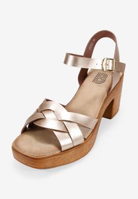 Metallic gold women's sandal with an open toe, crisscross straps, ankle buckle, and a chunky wooden heel. Textured leather and smooth footbed.