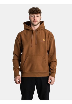 Sudadera marrón hecha de tela suave, con un bolsillo frontal, capucha con cordón y un pequeño logotipo en amarillo. El modelo está de pie frente a un fondo neutro.