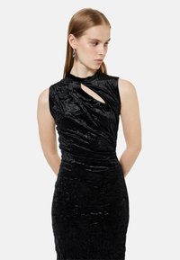 Sort ærmeløs kjole med høj hals og et snoet udsnitdesign. Stoffet har en tekstureret, krøllet overflade.