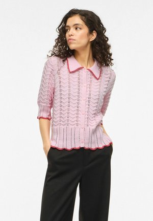 Kobieta o kręconych ciemnych włosach nosi różowy sweter na guziki z czerwonym wykończeniem i czarne spodnie, stoi z rękami w kieszeniach, patrząc w lewo.