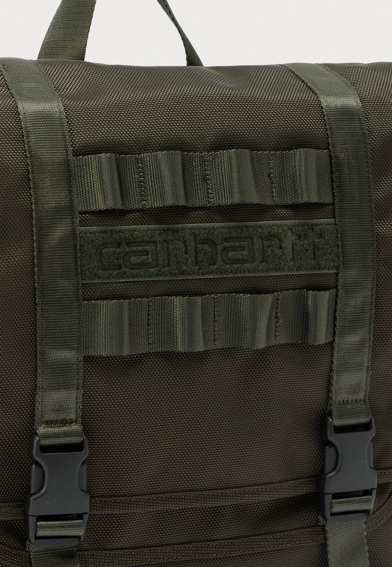 Sac à dos Carhartt vert foncé avec des bretelles robustes en sangle, des fermetures à boucle en plastique et une poignée de transport supérieure.