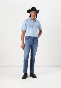 Lyseblå denimskjorte med korte ermer og to brystlommer, kombinert med blå jeans og svarte lærsko. Modellen har på seg en svart cowboyhatt.
