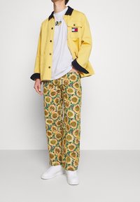 Žlutá bunda s námořnickým límcem, bílý tričko a kalhoty s vzorem slunečnic. Bílé tenisky dotvářejí outfit.