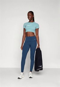 Bluză scurtă albastru deschis cu inscripția "Nike" și colanți înalți bleumarin. Modelul ține un prosop negru. Pantofi sport albi cu accente negre.