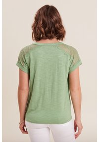 Grünes T-Shirt mit kurzen Ärmeln und Spitzenverzierungen an den Schultern, weicher Textur, runder Ausschnitt und lockerer Schnitt, kombiniert mit weißen Hosen.