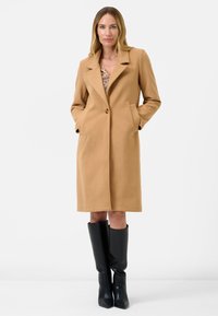 Camel-kleurige jas met een gestructureerd ontwerp, met inslagkleppen, een voorste knoop en zijzakken. Draagt over een gestreepte outfit en gecombineerd met zwarte laarzen.