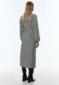 Schwarz-weiß gemustertes Kleid mit langen Ärmeln, tailliertem Bund und V-Ausschnitt auf der Rückseite, getragen mit kniehohen schwarzen Stiefeln.
